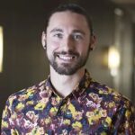 A man with a beard and long hair tied back, wearing a colorful floral shirt, stands indoors and smiles at the camera.