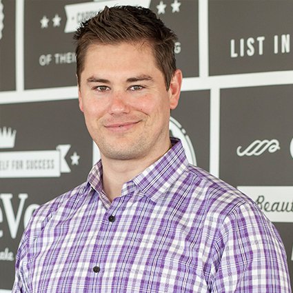 A man wearing a purple plaid shirt stands in front of a wall with various white text and graphic designs on a gray background.
