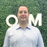 A man in a light blue dress shirt stands in front of a green hedge wall with large white letters "OM" partially visible behind him.