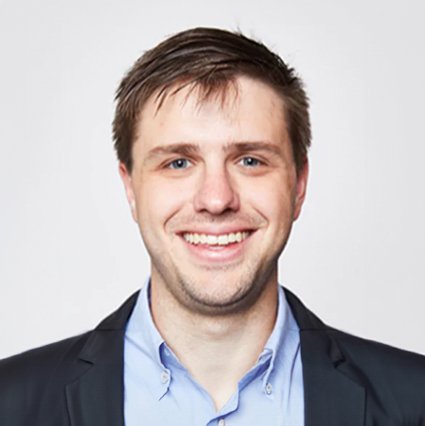 A man with short brown hair is smiling at the camera. He is wearing a dark blazer over a light blue collared shirt and is posed against a plain white background.