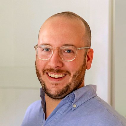 A smiling man with a bald head, glasses, short beard, and a light blue collared shirt stands in front of a neutral background.