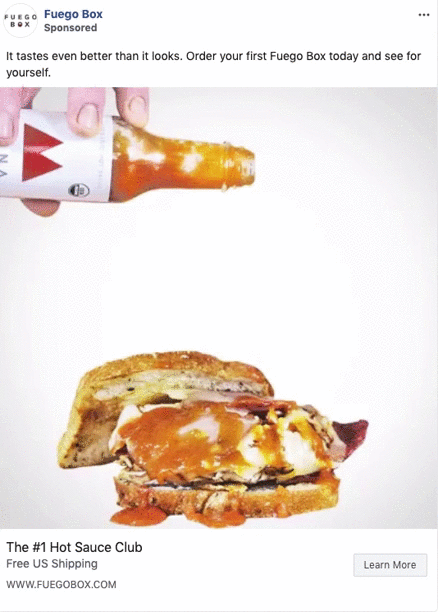 A hand pours hot sauce from a bottle onto a sandwich with meat, cheese, and sauce, placed on a white surface.