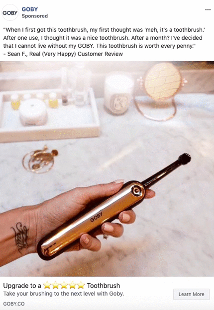 A hand holds a metallic electric toothbrush over a bathroom counter with grooming items, a lamp, and a round mirror in the background.