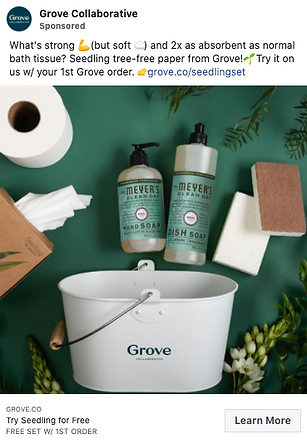 A white Grove bucket is displayed with Mrs. Meyer's soap bottles, a brown sponge, white towels, and green foliage on a marble surface.