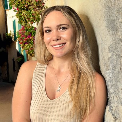 A woman with long blonde hair in a sleeveless beige top smiles while standing beside a stone wall and blooming flowers.