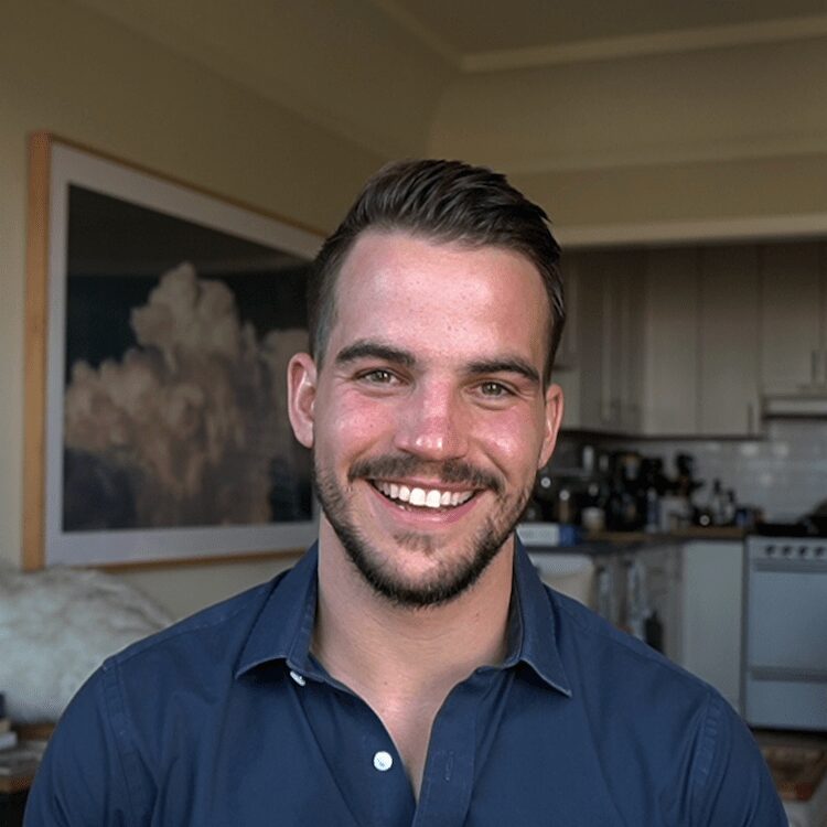 A man with short brown hair and a beard smiles at the camera, wearing a navy blue shirt, with a kitchen and large framed art in the background.
