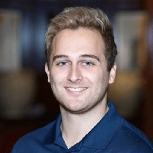 A young man with short blond hair and a trimmed beard, wearing a navy collared shirt, smiles at the camera against a blurred indoor background.