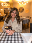 Woman with long brown hair sits at a checkered table in a restaurant, holding a glass of red wine and smiling at the camera.