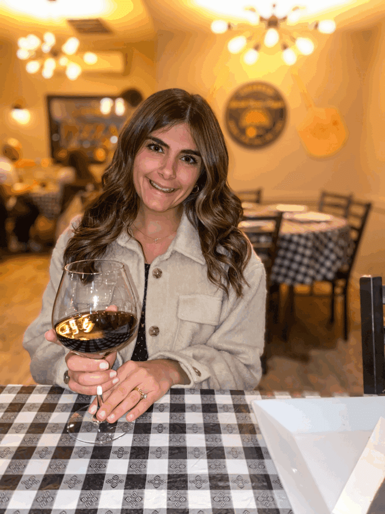 Woman with long brown hair sits at a checkered table in a restaurant, holding a glass of red wine and smiling at the camera.