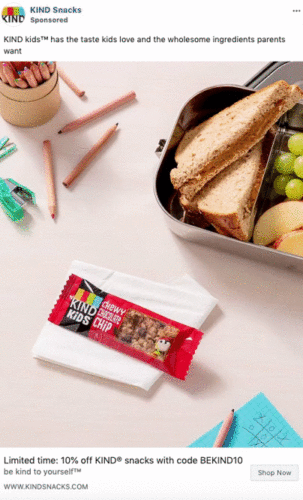 A KIND Kids chewy chocolate chip bar on a napkin next to a metal lunchbox with a sandwich, grapes, apple slices, and a pencil with a tic-tac-toe game.