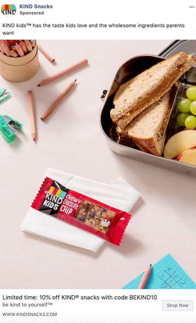A KIND Kids chewy chocolate chip bar on a napkin next to a metal lunchbox with a sandwich, grapes, apple slices, and a pencil with a tic-tac-toe game.