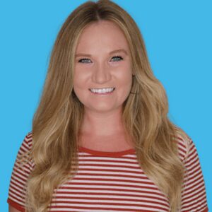 A woman with long blonde hair smiles at the camera. She is wearing a red and white striped shirt and is posed in front of a solid blue background.