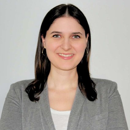 Woman with straight dark hair wearing a gray blazer and white top, standing against a plain light background, smiling at the camera.