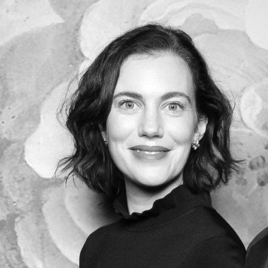 A woman with wavy dark hair, wearing a dark top with a ruffled collar, smiles at the camera in front of a patterned background. The photo is in black and white.