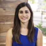 A woman with long brown hair wearing a blue sleeveless top and an infinity necklace stands indoors, smiling at the camera.