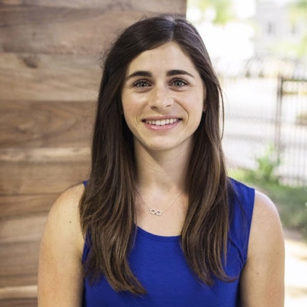 A woman with long brown hair wearing a blue sleeveless top and an infinity necklace stands indoors, smiling at the camera.