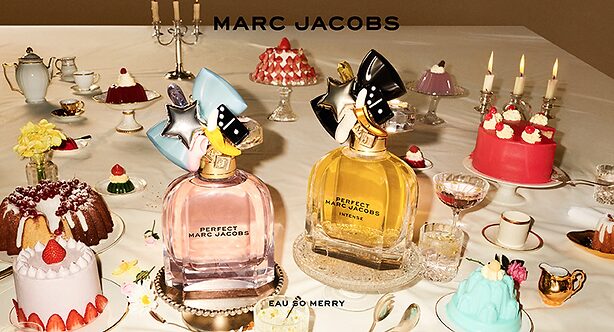 Two Marc Jacobs "Perfect" perfume bottles on a table surrounded by assorted cakes, pastries, candles, and tea sets.