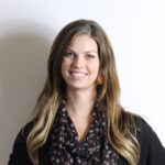 A woman with long brown hair wearing a black top, patterned scarf, and an earring stands in front of a plain white wall, smiling at the camera.