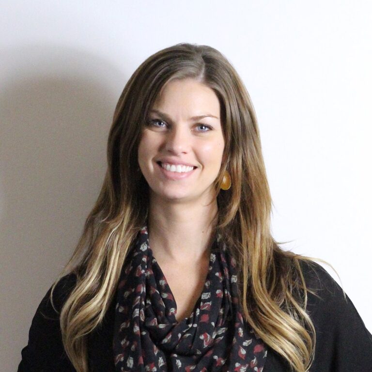 A woman with long brown hair wearing a black top, patterned scarf, and an earring stands in front of a plain white wall, smiling at the camera.