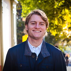 A young man with blond hair smiles at the camera outdoors, wearing a dark jacket over a collared shirt. Trees with green and yellow leaves are in the background.