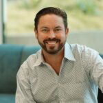 Man with dark hair and beard, wearing a patterned button-up shirt, sitting on a blue sofa and smiling at the camera.