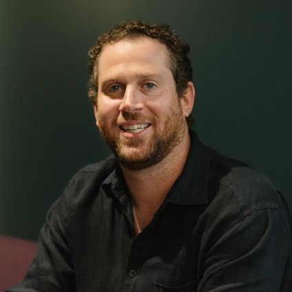 A man with short curly hair and a beard, wearing a dark button-up shirt, sits and smiles at the camera against a dark background.