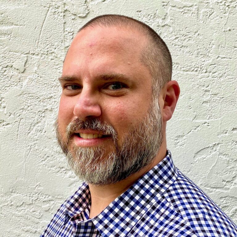 A man with a short beard and close-cropped hair, wearing a blue and white checkered shirt, stands in front of a textured white wall.