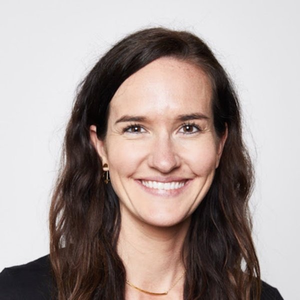 A woman with long brown hair smiles at the camera against a plain white background. She is wearing a black top, gold earrings, and a thin gold necklace.