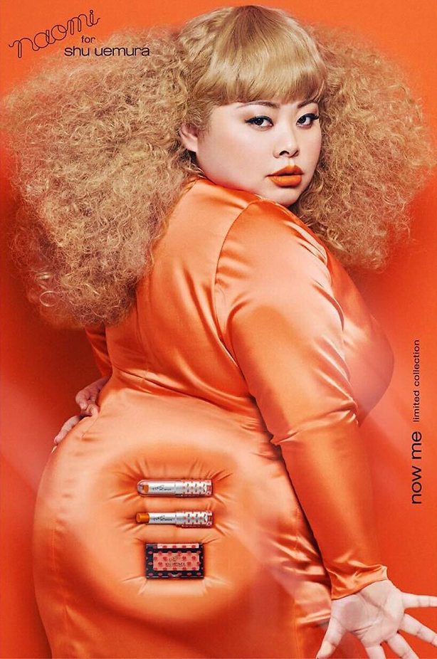 A woman with voluminous curly hair poses in an orange satin dress against an orange background, with cosmetics displayed in a pocket on her hip. Text reads "naomi for shu uemura" and "now me".