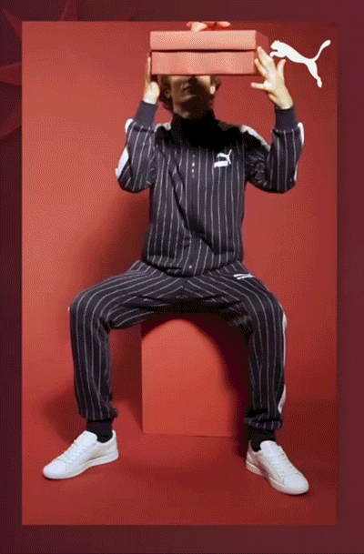 Person in a black pinstripe Puma tracksuit and white sneakers sits on a red block, holding an orange Puma shoe box above their head against a red background.