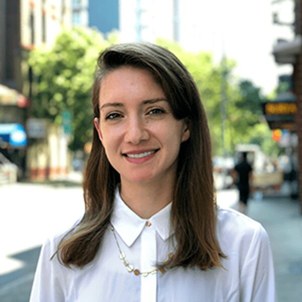 A woman with straight brown hair, wearing a white blouse and necklace, stands outdoors on a city street with buildings and trees in the background.