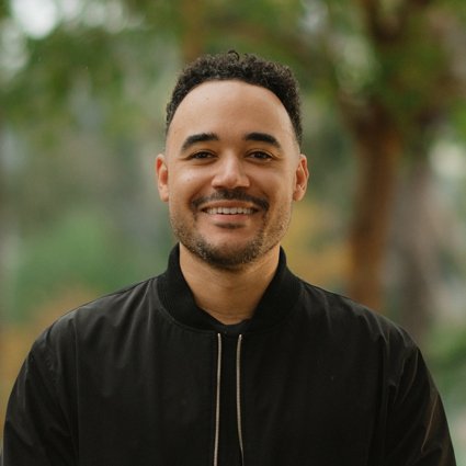 A man with short curly hair and a trimmed beard smiles at the camera, wearing a black jacket, with a blurred green outdoor background.