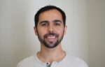A man with short dark hair and a trimmed beard is smiling, wearing a light gray henley shirt, standing in front of a plain light background.