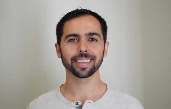 A man with short dark hair and a trimmed beard is smiling, wearing a light gray henley shirt, standing in front of a plain light background.