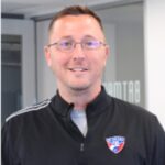 A man wearing glasses and a black jacket with a soccer team logo stands indoors, smiling at the camera.