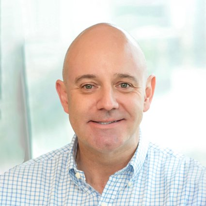 A middle-aged man with a shaved head, wearing a light blue checkered shirt, smiles at the camera in a bright indoor setting.