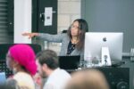 A woman gestures while speaking at the front of a classroom, with a computer and water bottle on the desk; several people are seated and listening.