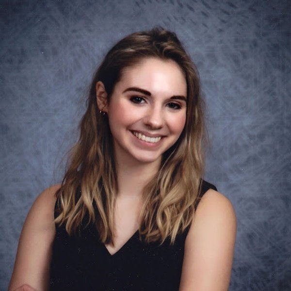 A woman with long, blonde hair wearing a sleeveless black top is smiling at the camera against a gray mottled background.