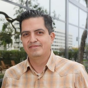 A man with short dark hair wearing a beige plaid shirt stands outdoors in front of a modern glass building and trees.