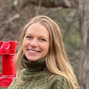 A woman with long blonde hair and a green sweater stands outdoors near a red metal structure, with trees in the background.
