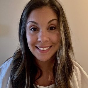 A woman with long brown hair and a white top smiles at the camera, standing in front of a plain beige background.