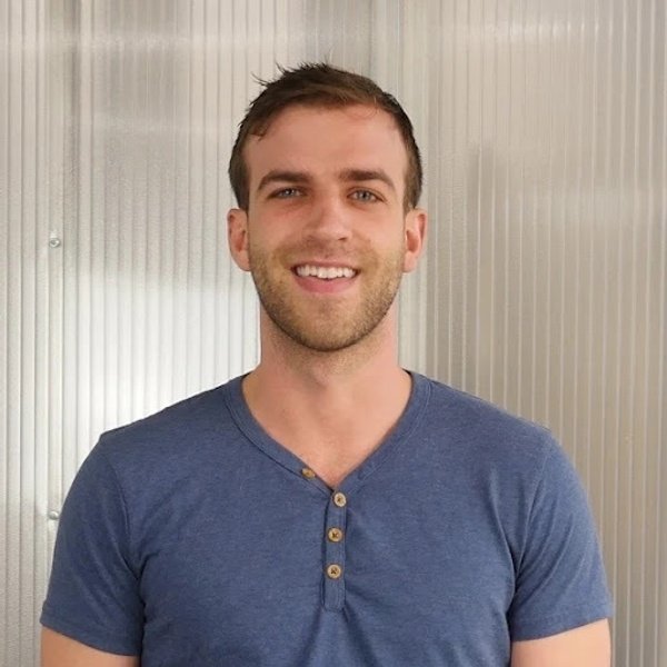 A man with short brown hair and a trimmed beard is smiling, standing in front of a corrugated metal wall, wearing a blue henley shirt.
