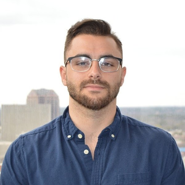 A man with short brown hair, a trimmed beard, and glasses wears a blue button-up shirt while standing in front of a cityscape background.