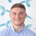 A man with short light brown hair smiles at the camera, wearing a light blue button-up shirt, with a white and teal bird-patterned background.