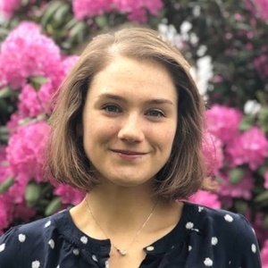 A woman with short brown hair and a navy polka-dot blouse stands in front of blooming pink flowers outdoors.