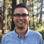 A man wearing glasses and a light blue button-up shirt smiles while standing outdoors in a wooded area.