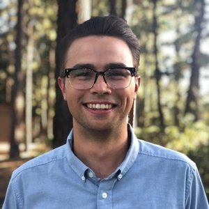 A man wearing glasses and a light blue button-up shirt smiles while standing outdoors in a wooded area.