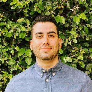 A man with dark hair and a trimmed beard wearing a blue button-up shirt stands in front of a leafy green background.
