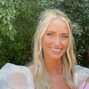 A woman with long blonde hair and a white dress stands outdoors in front of green foliage, smiling at the camera.