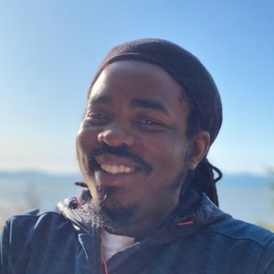 A man with a beard and mustache, wearing a dark head covering and jacket, smiles outdoors with a blurred sky and water in the background.